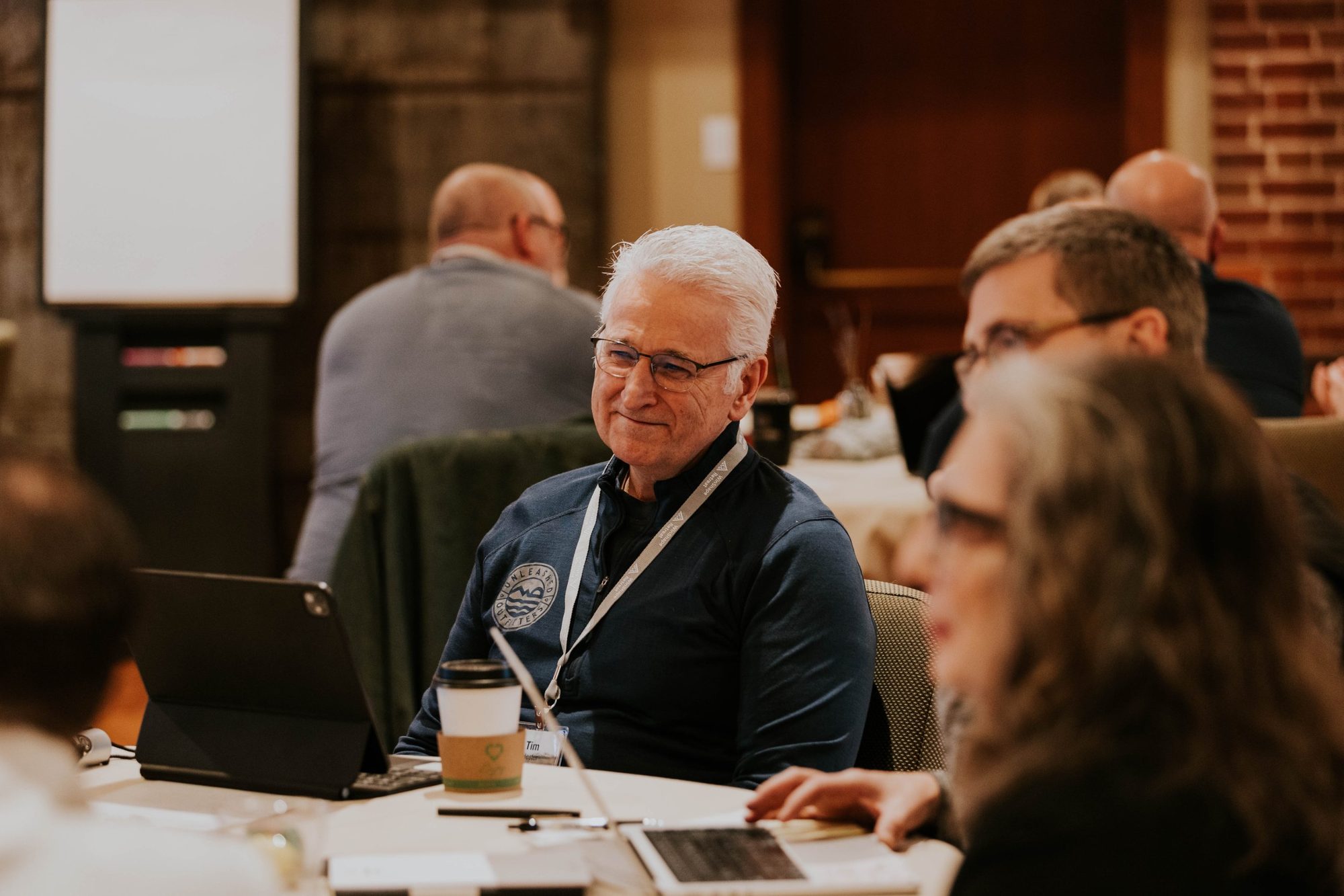 Veteran pastor laughing with peers during a WinShape Leadership Collective session, name tag visible, warm room setting