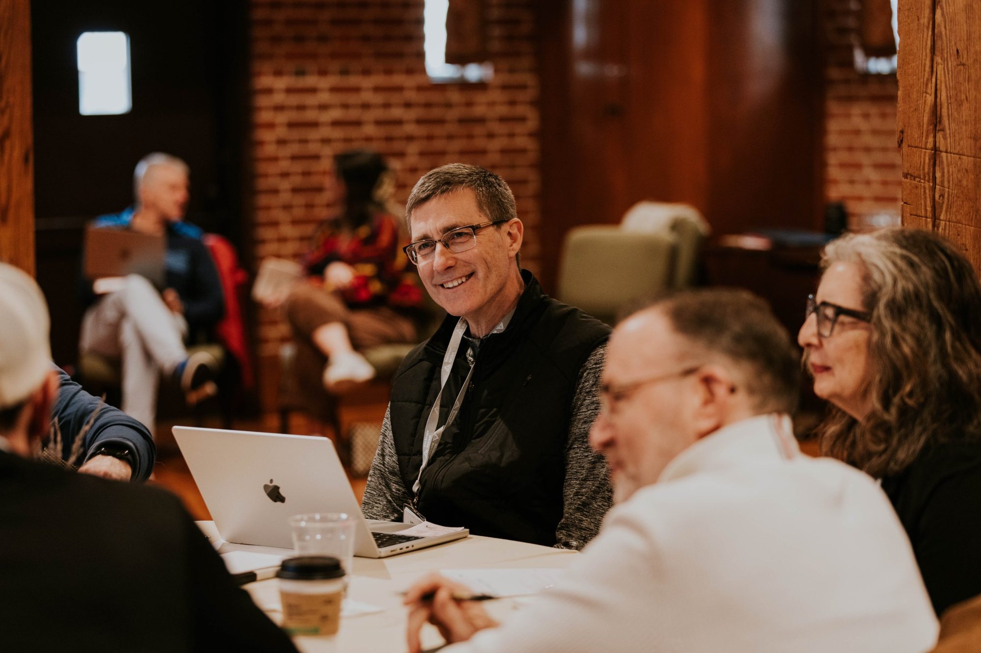Executive pastor smiling and engaging with peers at a WinShape Leadership Collective table discussion, laptop open, brick wall background